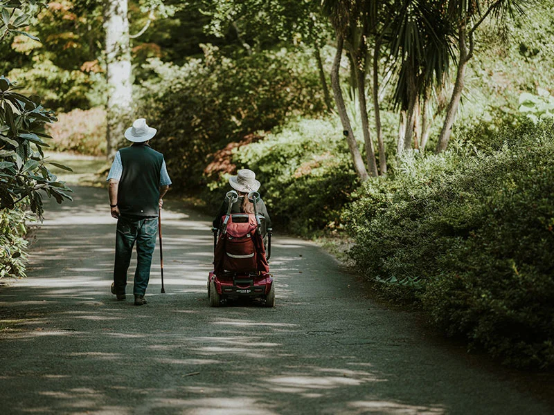 Two travelers on a shaded garden path, one using a mobility scooter